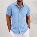 Mann in hellblauem Leinenhemd und weißen Shorts vor neutralem Hintergrund. Sommermode, Herrenbekleidung, lässiger Stil.