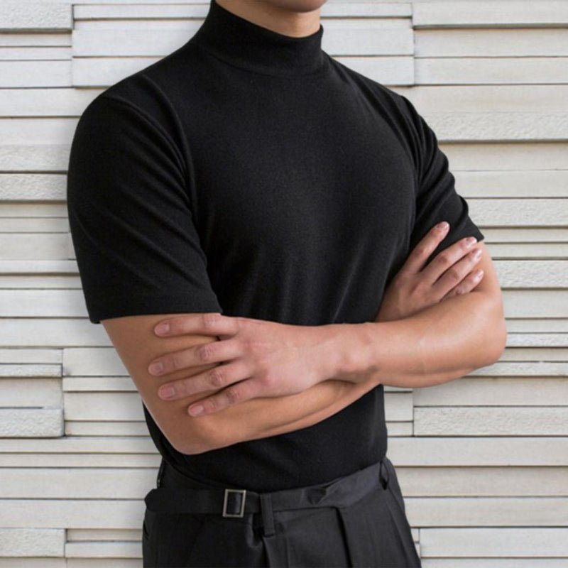 Mann in schwarzem Rollkragenpullover vor weißer Ziegelwand, Arme verschränkt. Elegante Herrenmode, minimalistischer Stil, zeitloses Design.